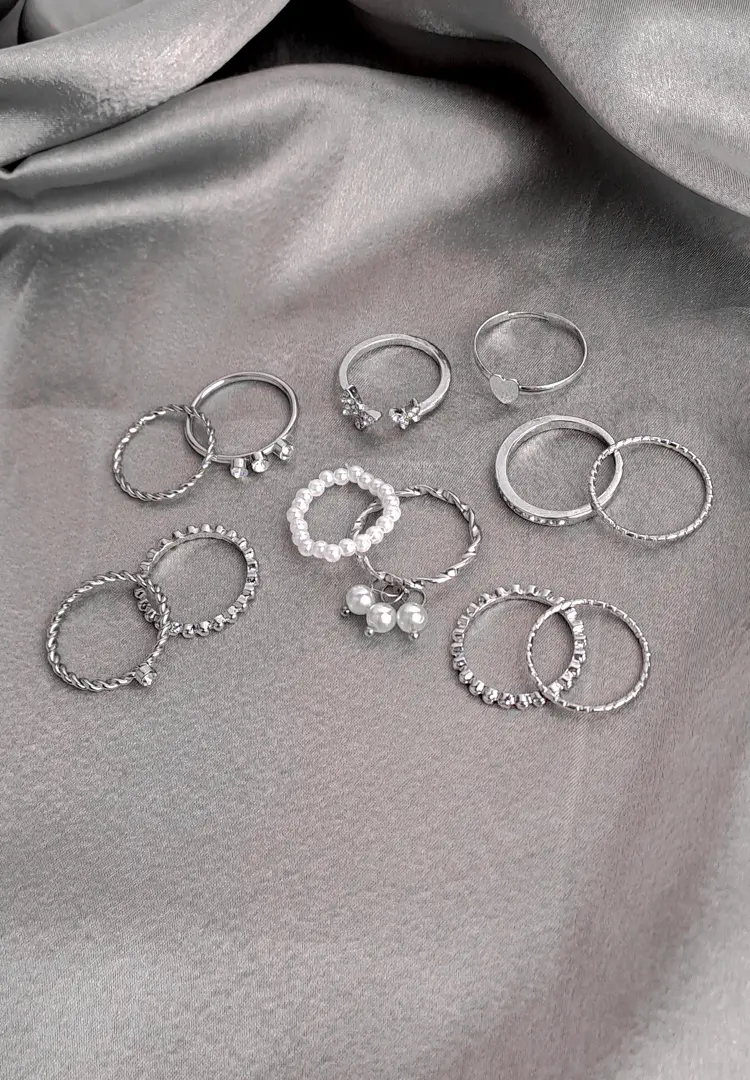 th (99) A large collection of twelve silver-toned stacking rings, including bands with pearl drops, continuous faux pearls, twisted rope texture, single crystal accents, and plain polished metal, scattered on a dark surface.