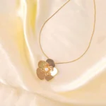 A gold-colored necklace featuring a large, five-petal flower pendant with a white faux pearl at its center and textured gold details