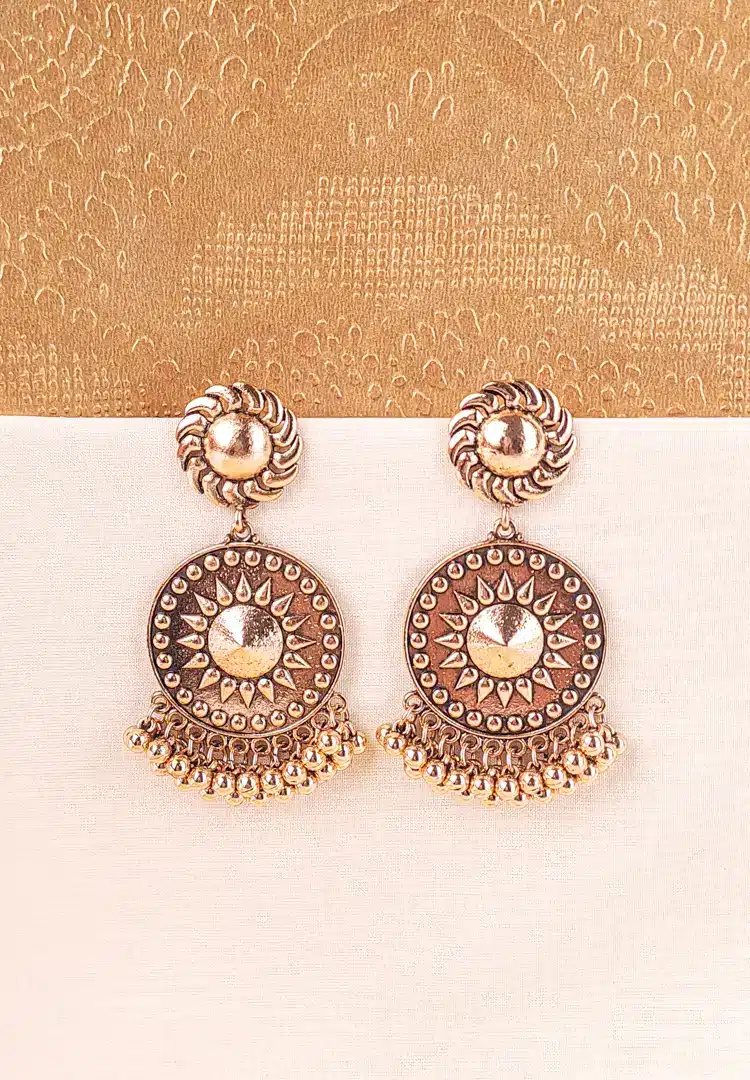 th - 2025-12-12T133631.967 Large, antique matte gold-tone drop earring featuring a circular disc with a raised sunburst or Chakra motif in the center, hanging from a swirling stud. The bottom is fringed with small, round ghungroo beads.