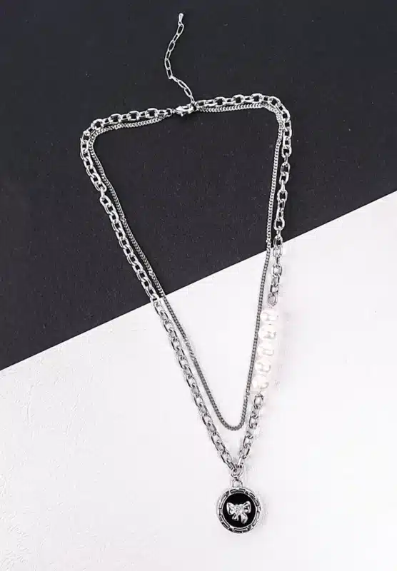 A silver-toned layered necklace is displayed on a dark gray laptop keyboard. The necklace features two main strands: one is a chunky curb chain with a circular pendant depicting a silver teddy bear, and the other is a finer chain adorned with five evenly spaced, lustrous white pearls. Both chains connect at the clasp end.