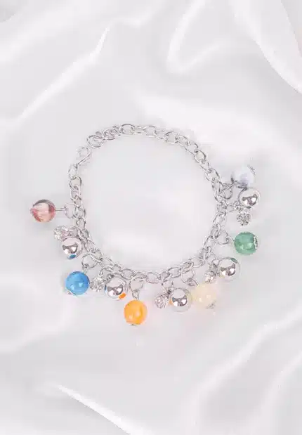 The bracelet features a robust chain from which various round charms dangle. These charms include polished, spherical beads in a range of colors such as turquoise, orange, a pale yellow-green, and a mottled white, interspersed with smaller, shiny silver spheres. The bracelet is clasped, forming a gentle curve