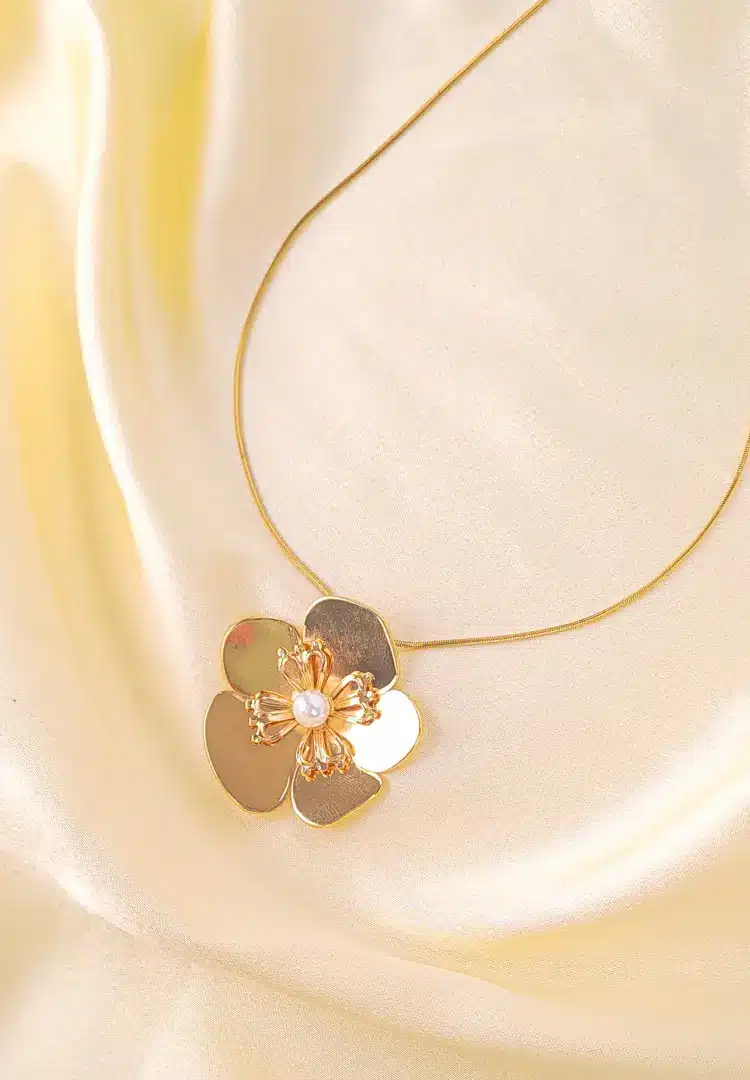 th - 2025-12-12T173349.652 A gold-colored necklace featuring a large, five-petal flower pendant with a white faux pearl at its center and textured gold details