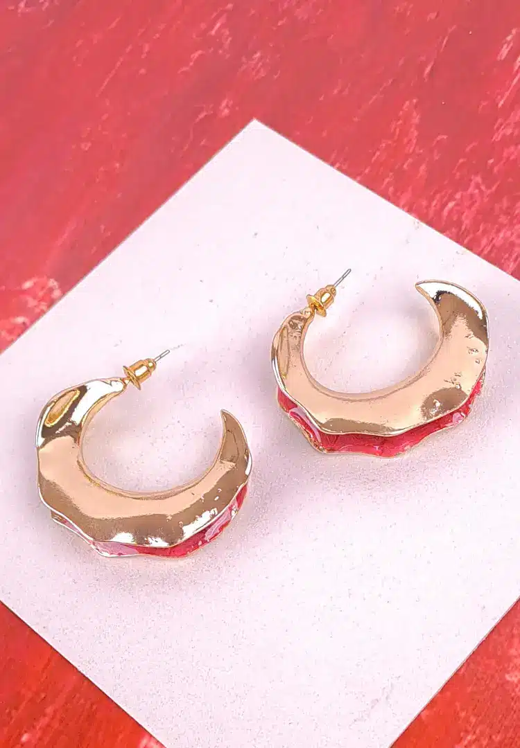 th - 2025-12-12T181314.321 The earrings are gold-toned and feature an organic, undulating shape, with one side of each hoop partially coated in a glossy, rich crimson or deep red enamel.